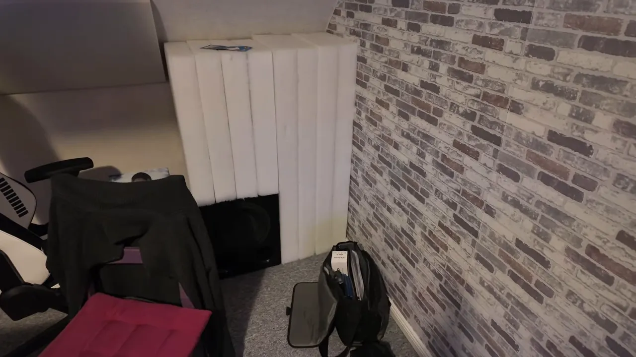 Weiß verkleideter Bassabsorber in der hinteren Ecke unter der Dachschräge mit einem schwarzen Subwoofer im unteren Bereich und Bürostuhl links